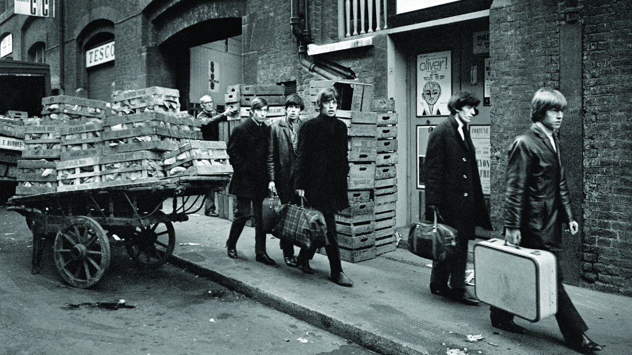 The Rolling Stones walking past a market trader down a Soho street carrying suitcases.