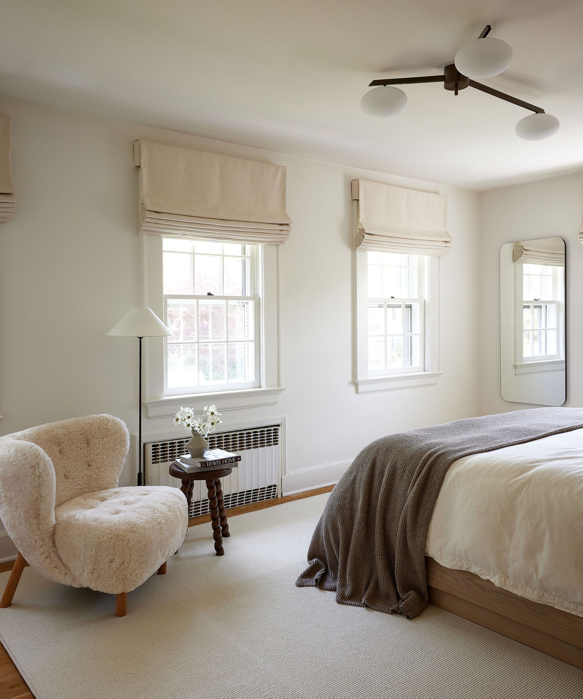 white bedroom with traditional windows with cream Roman blinds, neutral bedding, a light beige rug, and a modern cream accent chair