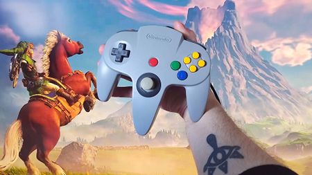 Hand holding grey N64 controller in front of Zelda Ocarina of Time gameplay trailer on monitor with Link on Epona looking and Death Mountain in the distance.