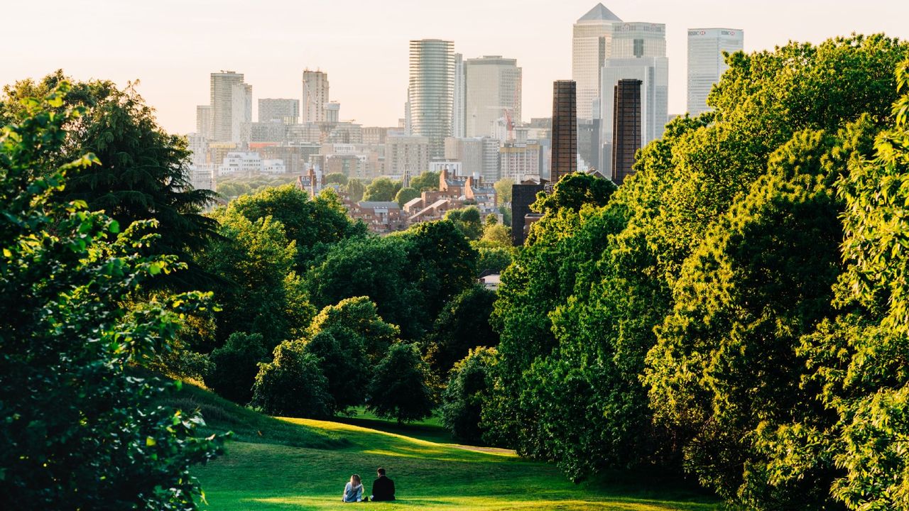 Two people sit in a tree lined park in front of a city skyline