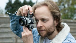 Man wearing blue denim jacket holding Nikon FM and looking through the viewfinder