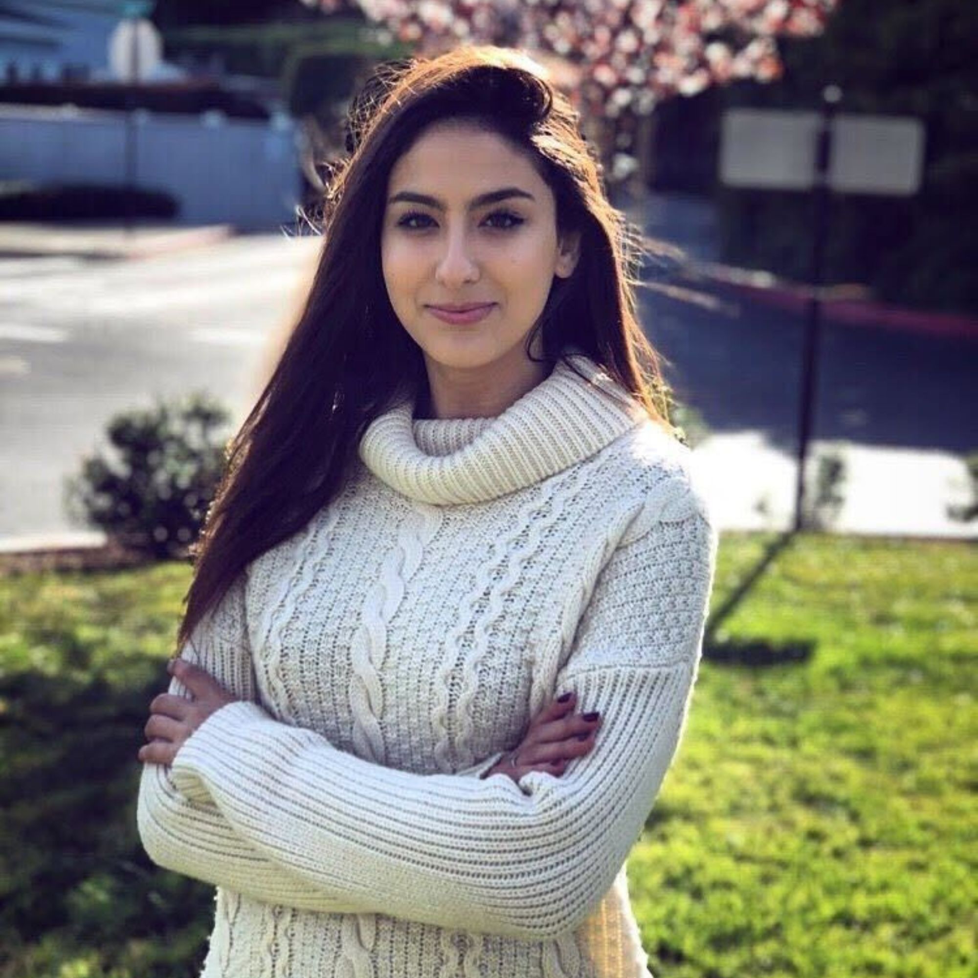 A picture of Malak Bellajdel, a woman with long brown hair wearing a white jumper with her arms folded, in front of a road with a lawn and a cherry blossom tree