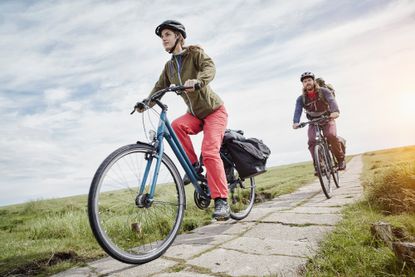 Best women's hybrid bikes main image is of a women riding a bike on a path between two fields with man on a hybrid bike behind her