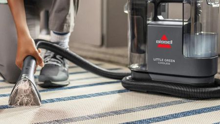 An olive-skinned female using the Bissell Little Green Cordless Portable Carpet Cleaner on cream and blue-striped area mat