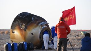 a scorched cone-shaped capsule lies on a desert floor surrounded by people in thick blue coats