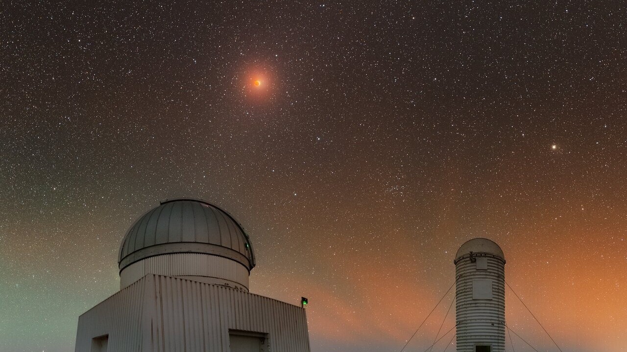 Two domed observatories sit underneath a green and orange night sky where a lunar eclipse shines down