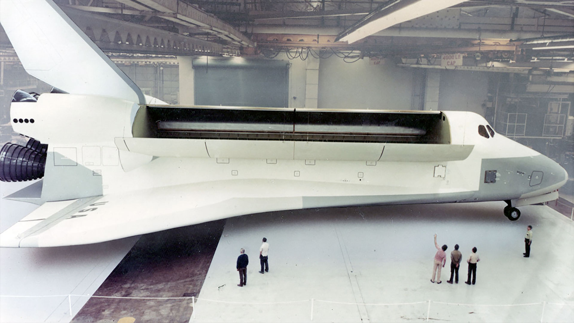 a full-size, all-white space shuttle model sits inside a hangar, with six people looking at it