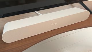 Sonos Ray in white on a wooden TV stand in front of a Sony TV