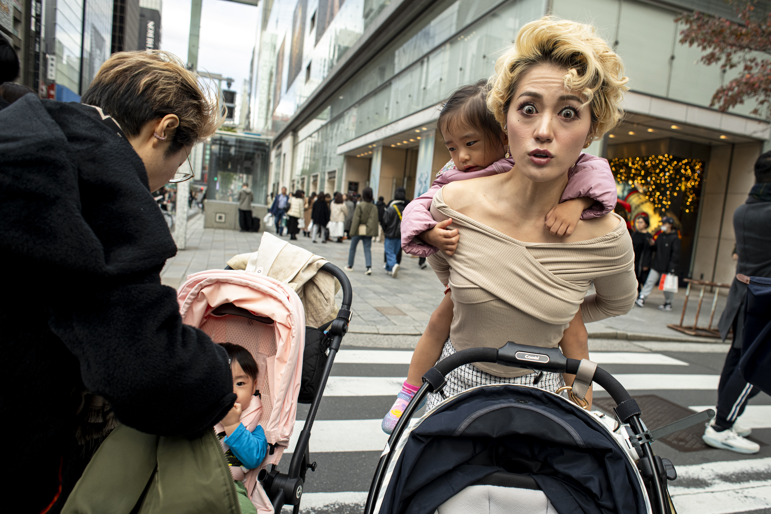 A woman with blonde hair and a surprised expression crosses a busy city street while carrying a young child on her back.