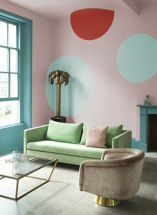 A living room paint color idea with Pashmina pink walls and circles of accent colors by Crown, with a green sofa and a beige accent chair, and a glass coffee table by the window