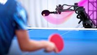 A black arm with a ovular paddle on the end passes a ball forward on a blue table tennis to a person with a red paddle in the foreground