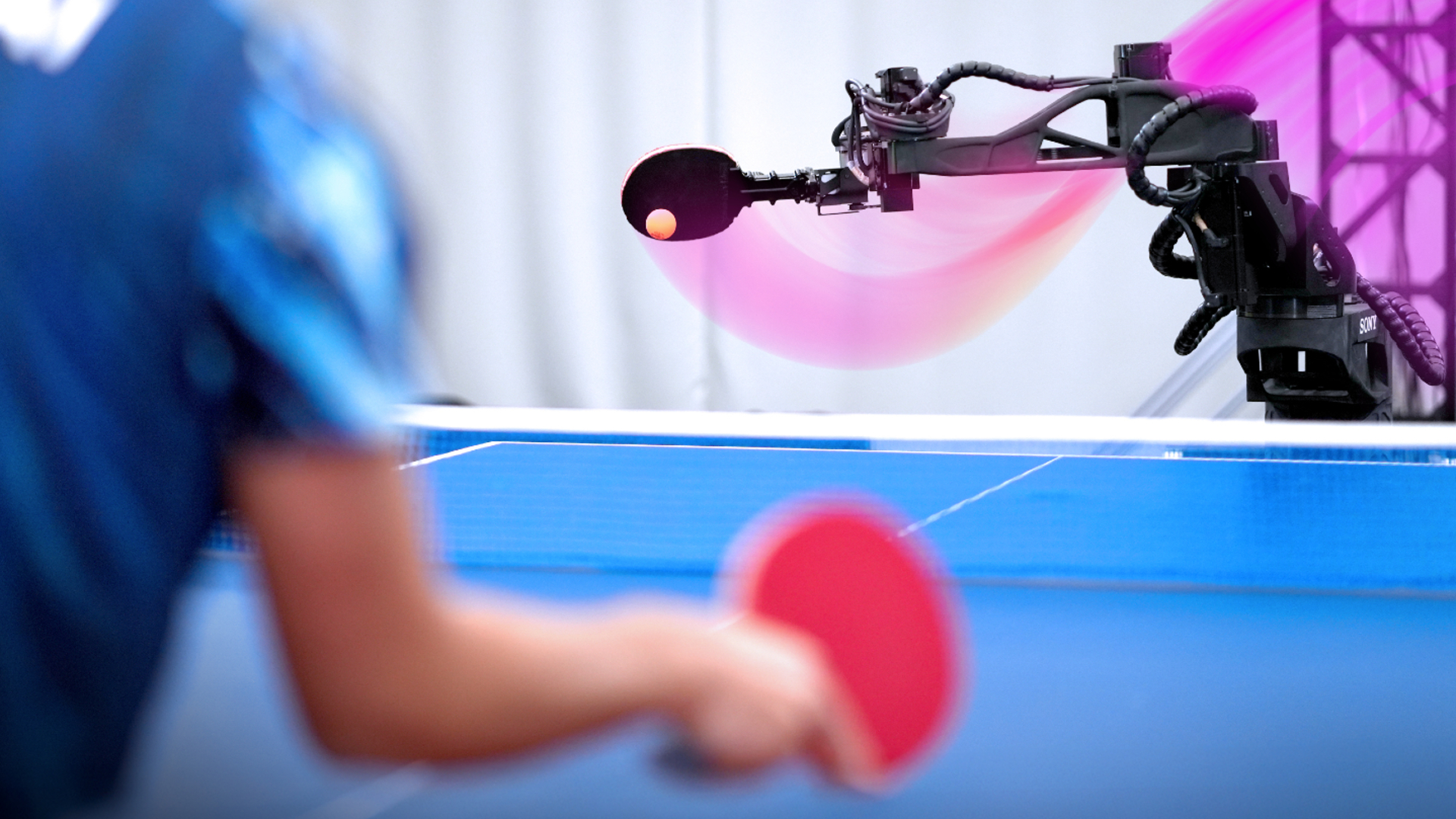 A black arm with a ovular paddle on the end passes a ball forward on a blue table tennis to a person with a red paddle in the foreground
