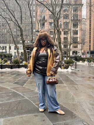 Ashlyn Delaney wearing a brown leather jacket, black leather tube top, jeans, furry hat, brown suede sneakers, and brown bag