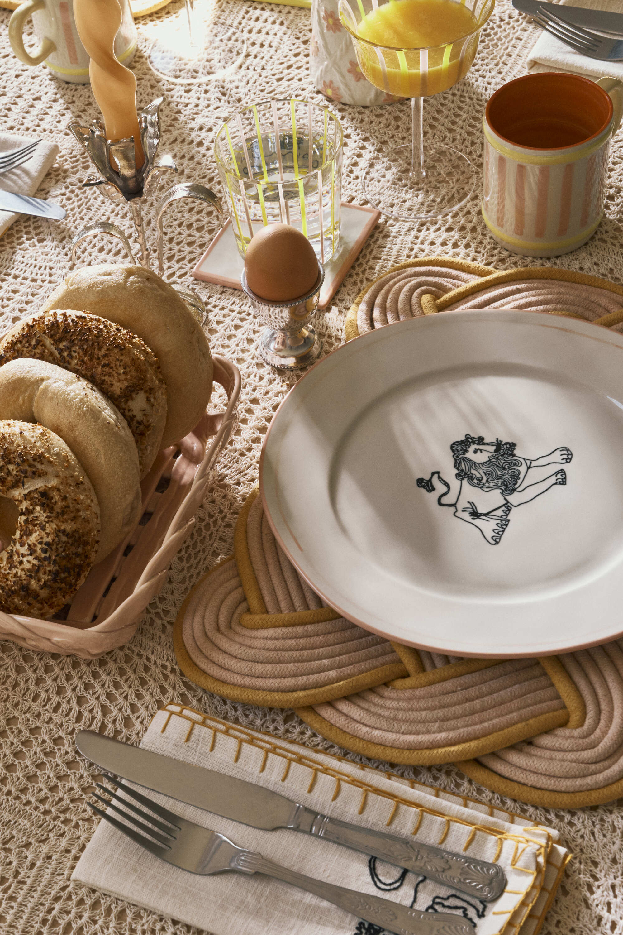 Image of a place setting at a brunch table. There is a crochet tablecloth, a pink and orange woven place mat and a ceramic plate with a lion on it.