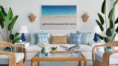 Nautical living room with coastal picture, potted palms, cream sofa with blue pillows, glass coffee table, and rattan chairs