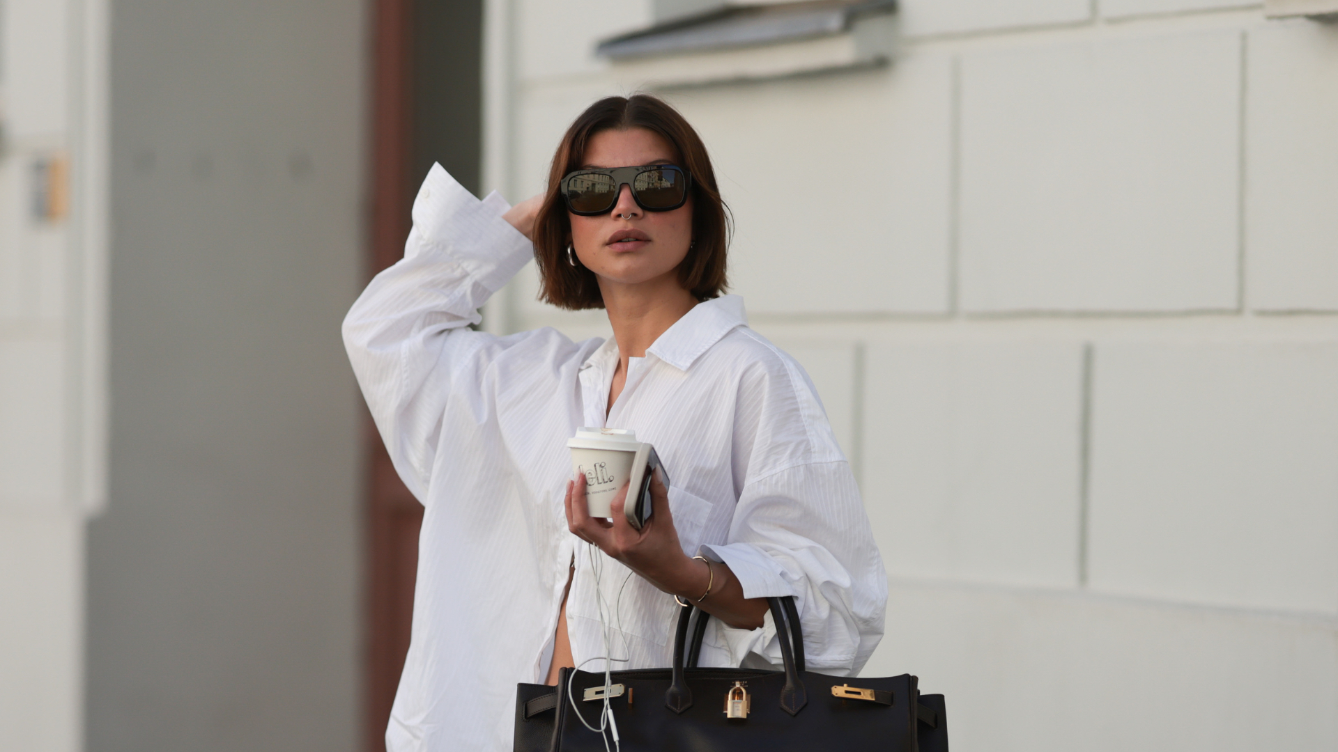 Sophia Geiss is seen wearing chunky silver hoop earrings, a silver nose ring, black aviator sunglasses from Vehla Eyewear, an oversized white structured shirt from Black Palms, wide-leg blue-grey Bermuda shorts from Drykorn, a large rectangular black leather “Birkin” bag from Hermès, a gold bangle, and black flip-flops from Havaianas, while holding a coffee cup and an Apple iPhone paired with Apple Wired EarPods during Berlin Fashion Week SS26 on June 30, 2025 in Berlin, Germany