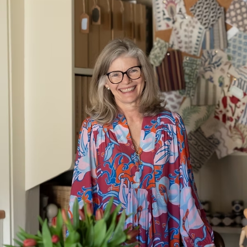 Image of a woman in a pink and blue floral-patterned dress and glasses. She is standing in a butter yellow room, and there are fabric samples on the wall.