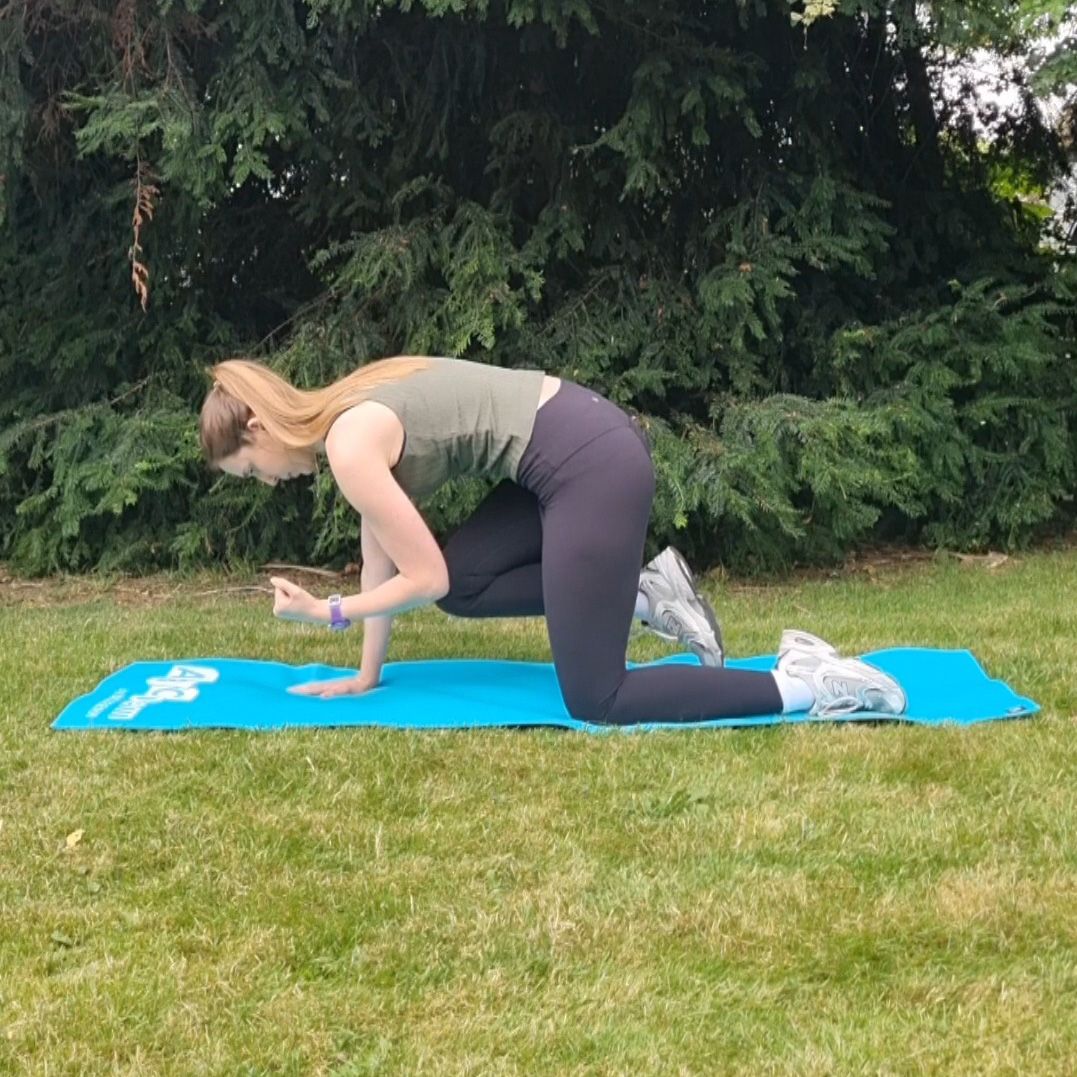 Fitness editor Ruth Gaukrodger performs a bird dog exercise in an outdoor setting on a yoga mat. Her left knee and shin lie on the yoga mat, and her right hand is on the ground. Her back is bent slightly as she crunches inwards, bringing her left elbow and right knee into contact below her torso. Behind her we see a pine tree.