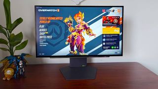LG UltraGear 27GX790A-B gaming monitor on woodgrain desk next to plant, Pharah figure, Samus toy, and Bigfoot Squishville with Overwatch 2 main menu on screen featuring Juno.