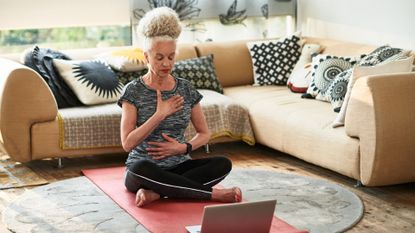 A woman does breathing exercises in her living room. She is sat on the floor on a yoga mat in front of a corner couch. One hand is on her chest, one on her belly and her legs are crossed.