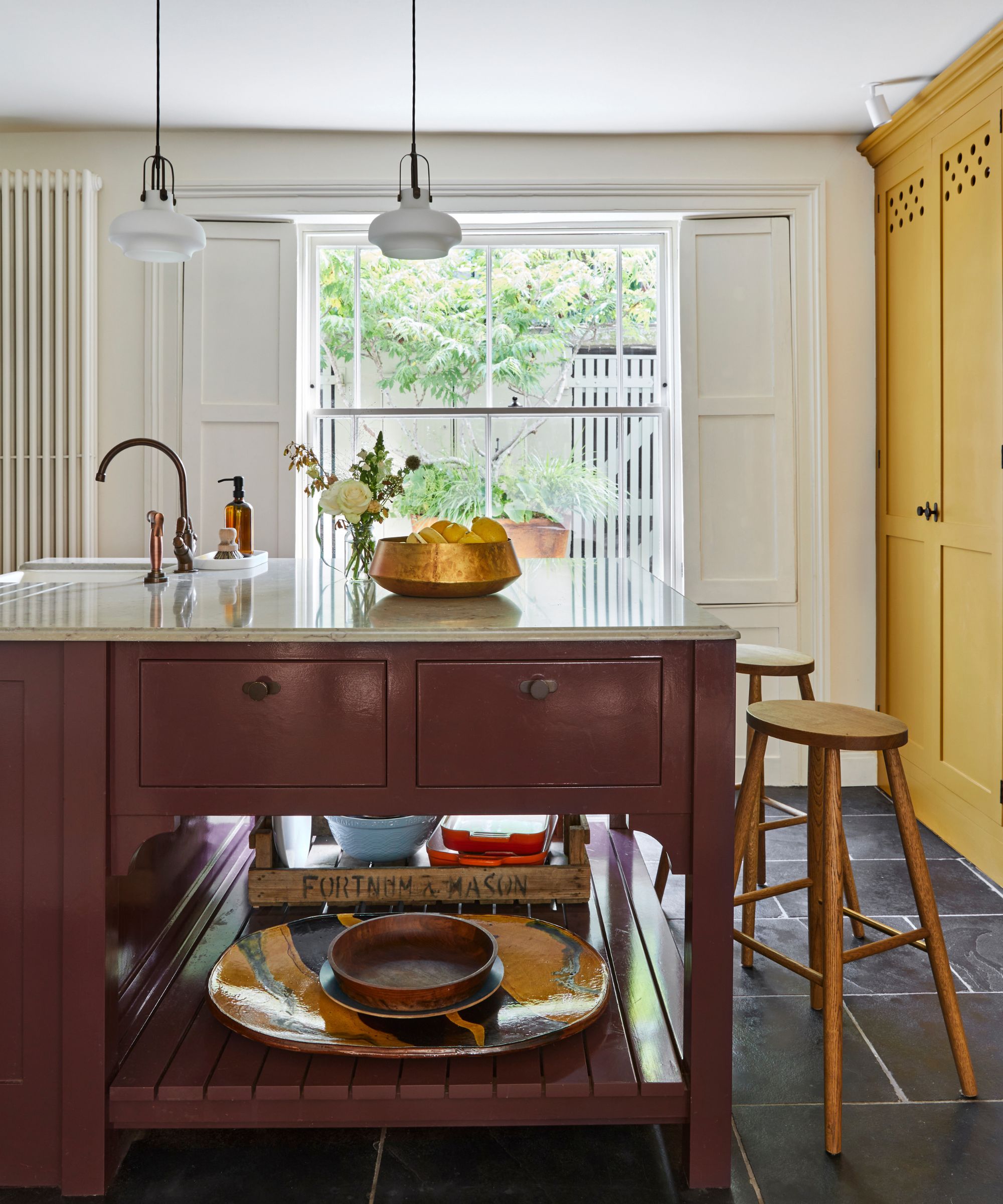 a kitchen with a freestanding island painted burgundy with decor on the bottom shelf and decor on the countertop, two stools to the right, and a yellow cabinet on the right side