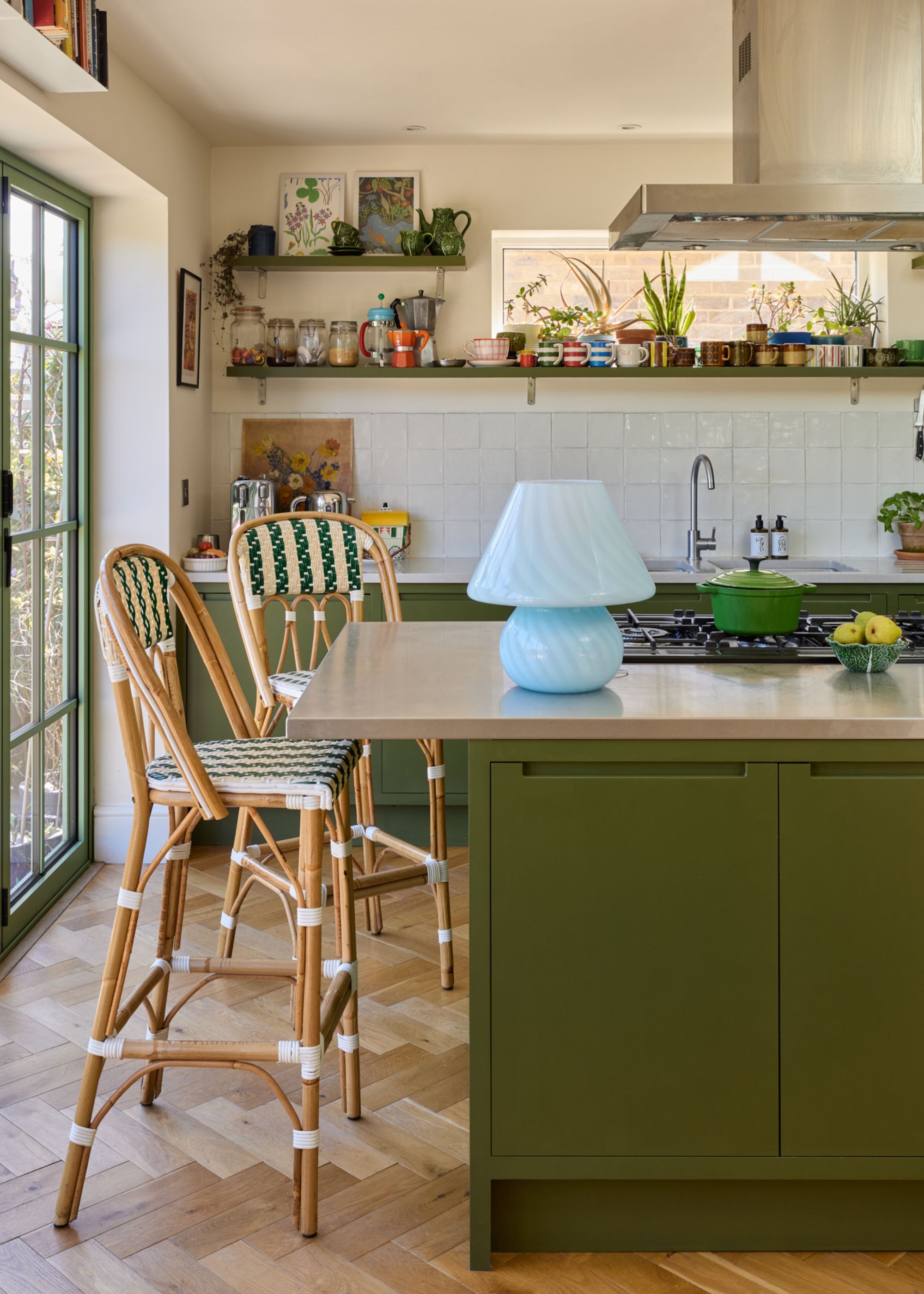 A dining kitchen with olive green cabinets and a kitchen island with a stainless steel counter, warm white walls, parquet flooring, and two bamboo bar chairs.