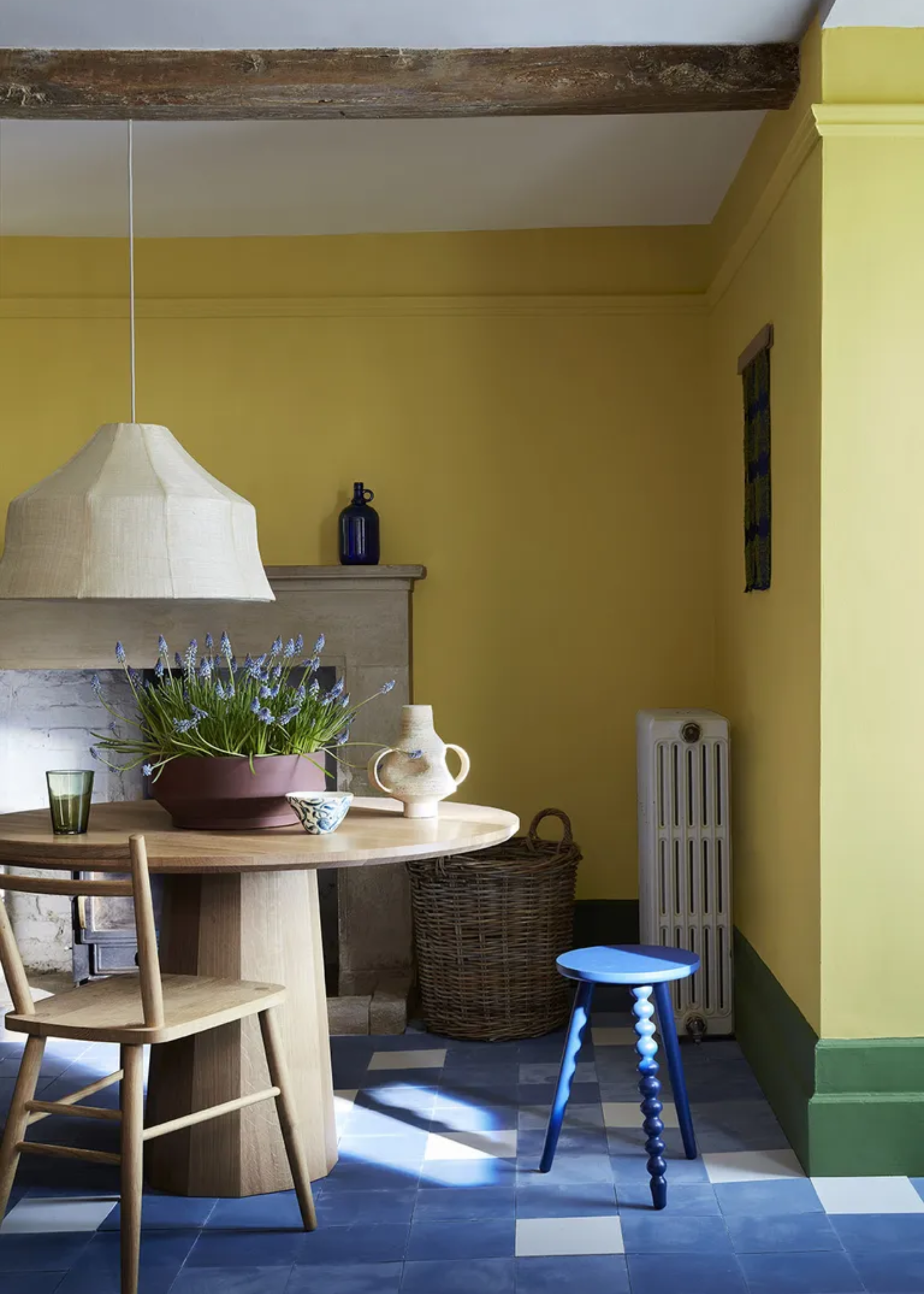 A yellow dining room with a blue tiled floor, a blue stool, and a wood dining table with potted lavender