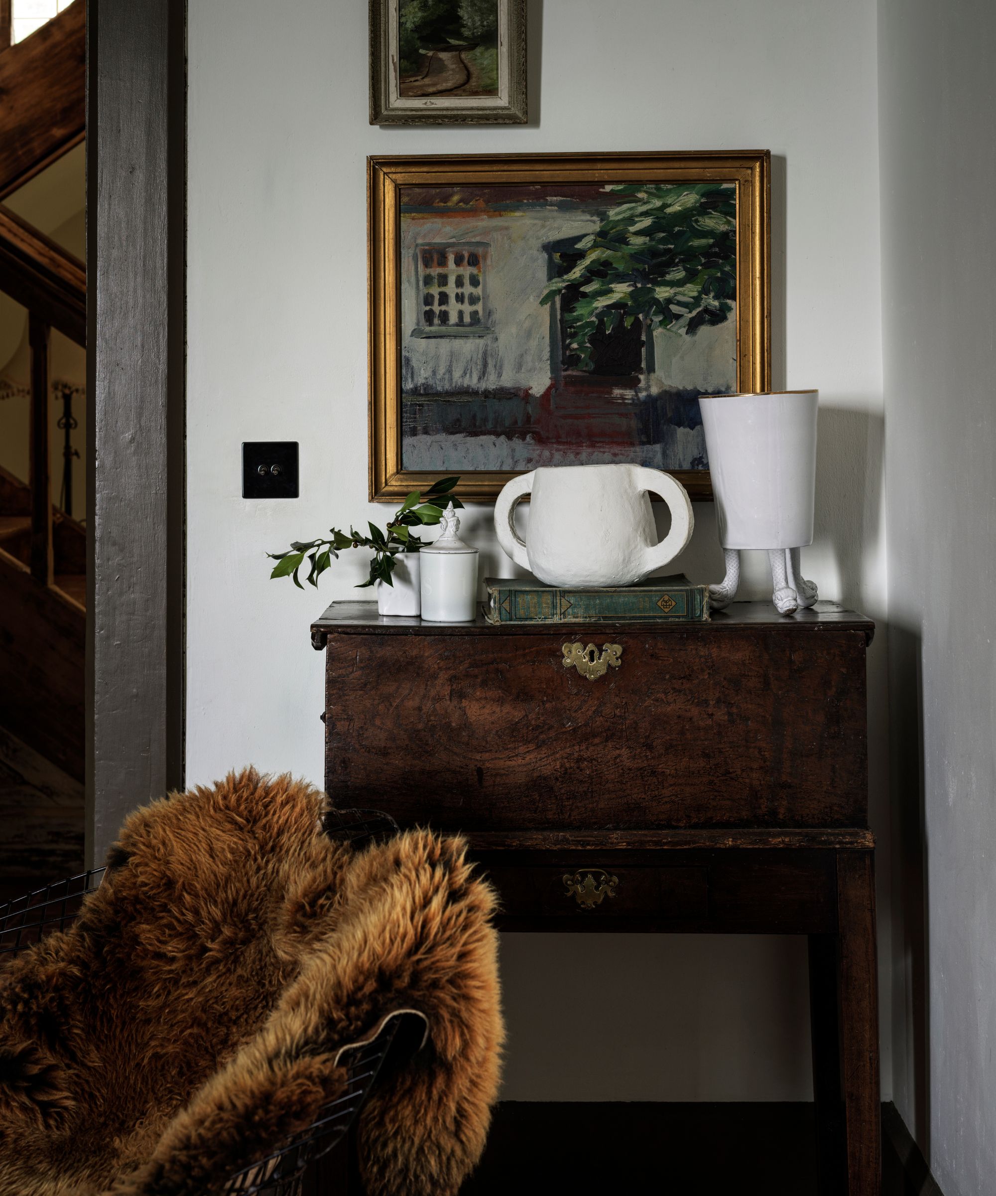 Mahogany chest of drawers with white porcelain ceramics on top, antique paintings hanging on the wall above, and a chair with a draped fur throw over it.