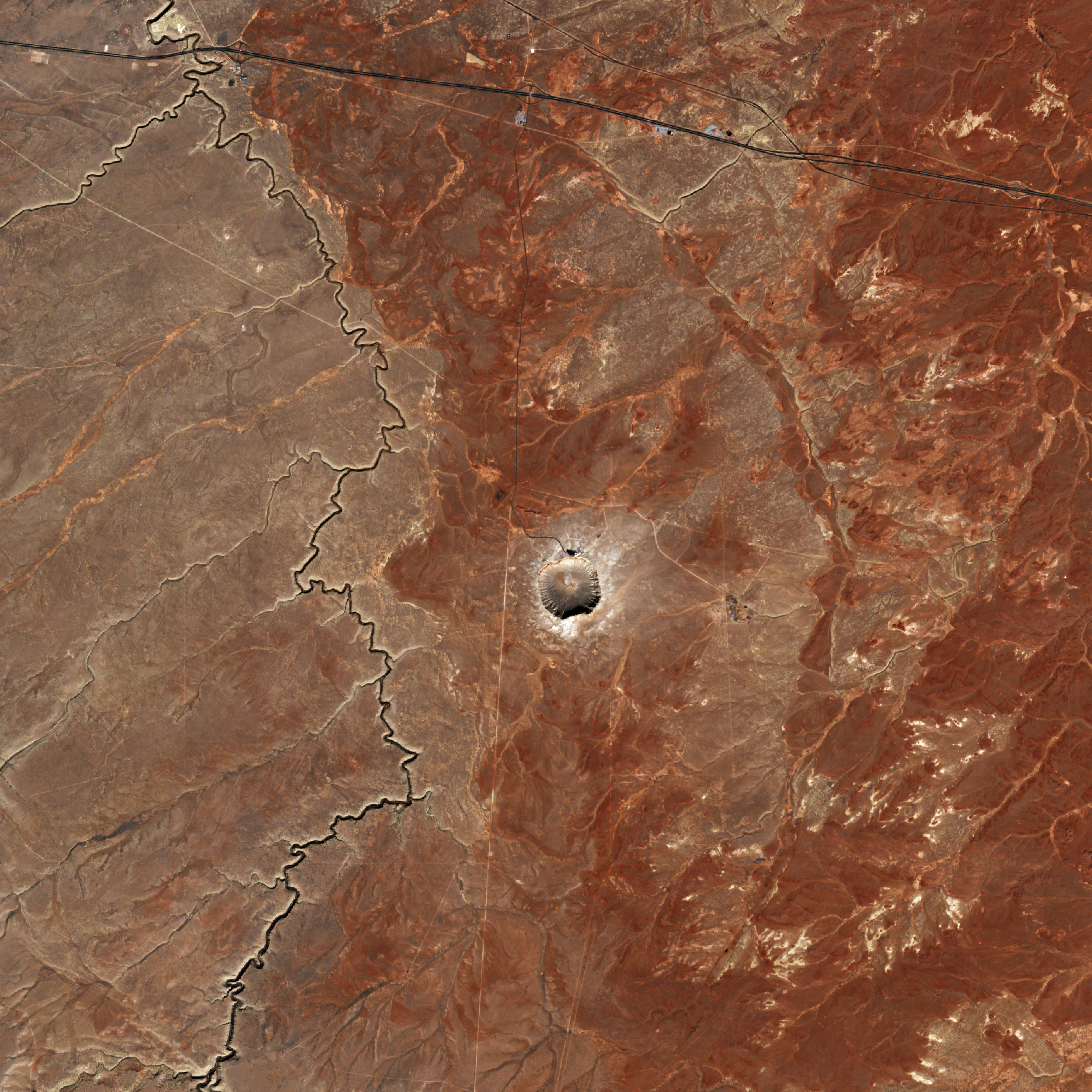 A small, relative to the image, crater lays in the center, the sunlight casting a short shadow from its rim upward into its bowl. The crater is surrounded by a barren rusted landscape with a thin jagged river on the left, , and the smooth line of a paved road sloping gently downward from the top left corner to just below the top right.