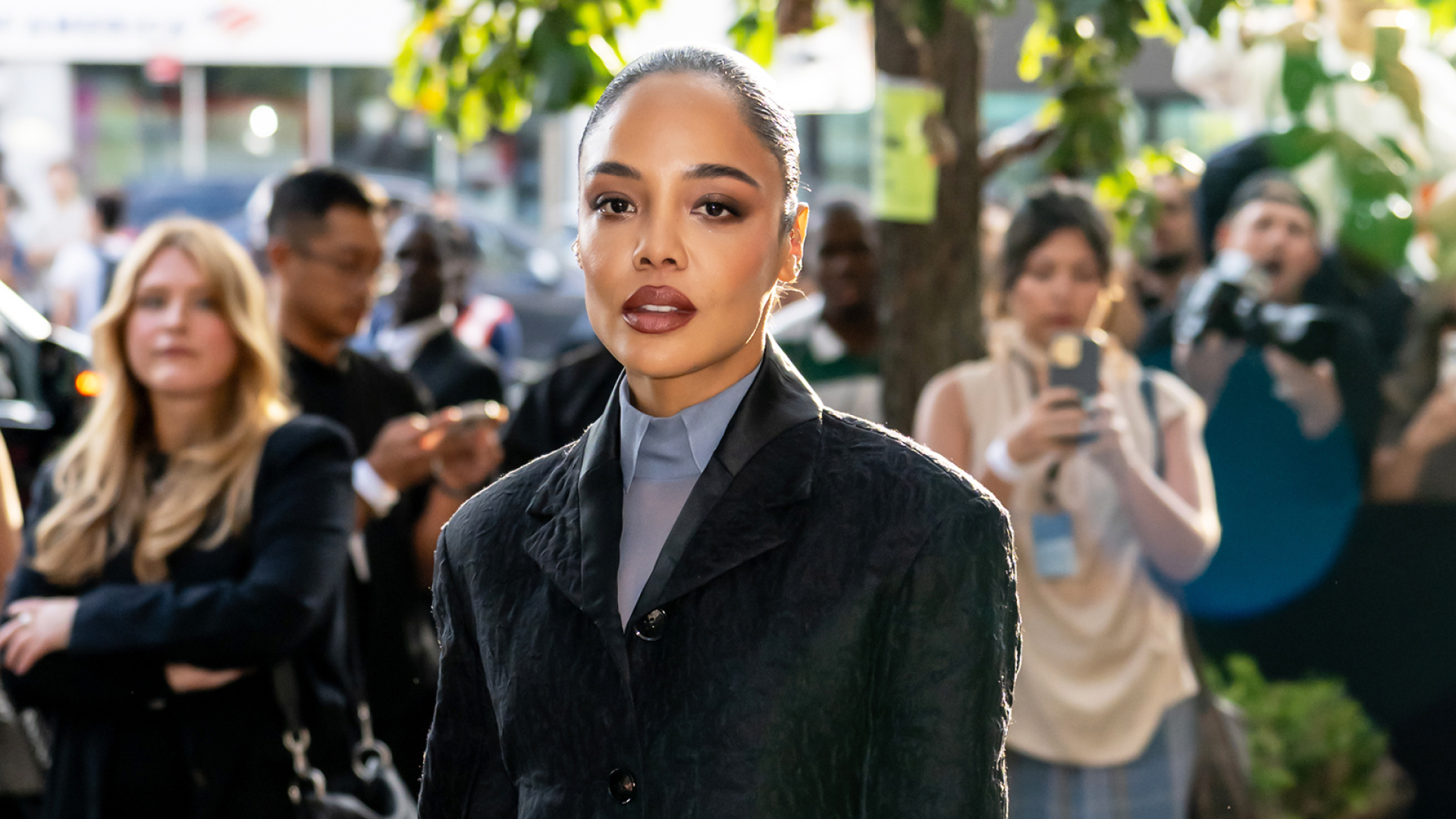 Tessa Thompson wearing a slicked-back bun hairstyle, a blazer, a button up shirt and dark pink lipstick.