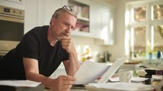 An older man peruses paperwork, looking thoughtful, while standing at his kitchen counter.