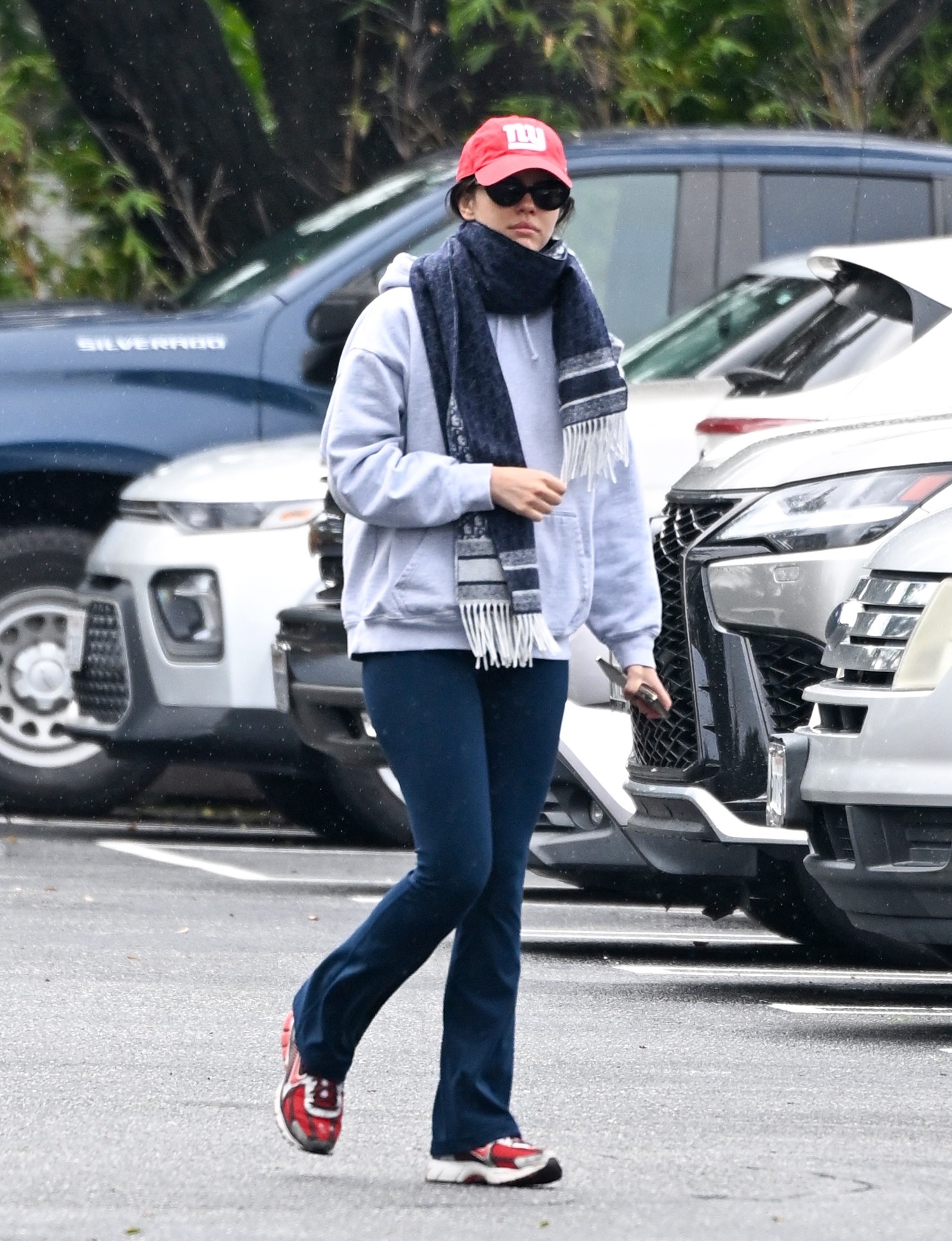 An image of Mia Goth wearing navy flare leggings with a white grey hoodie, red cap and red trainers.