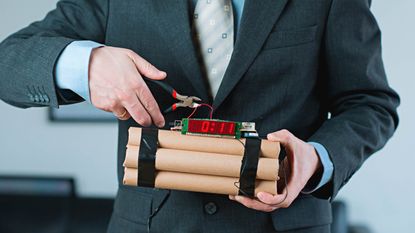 A man in a suit clips a wire on top of a stack of dynamite to defuse a bomb.