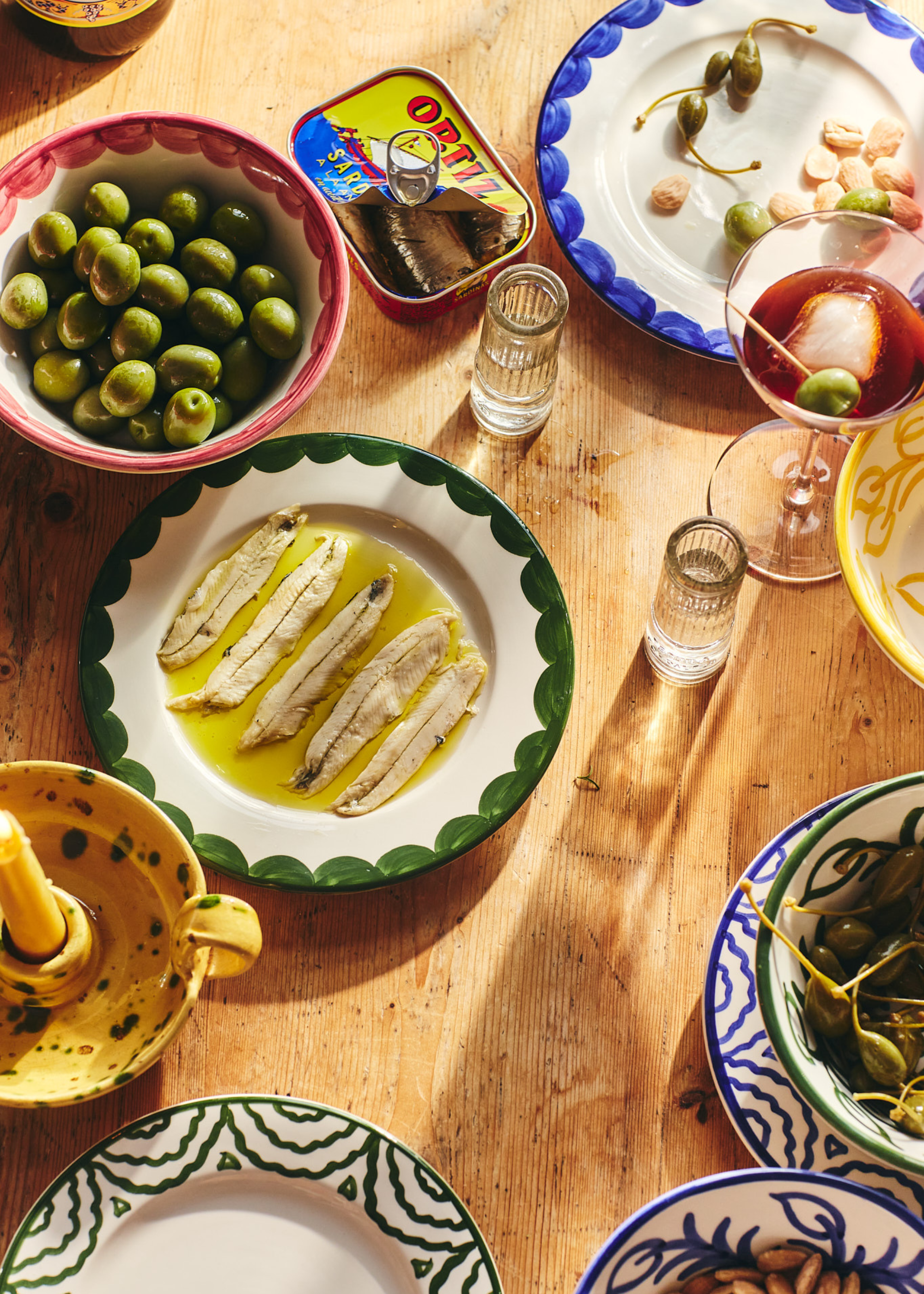 A table with hand-painted plates holding olives, tinned fish, nuts and bites alongside shot glasses, drink glasses and a splattered candle holder