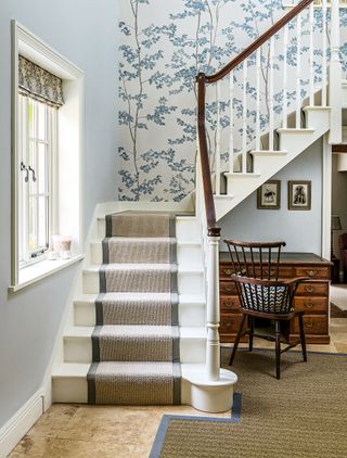 staircase with window and neutral runner and blue and white floral wallpaper dark wood chair and desk under stairs