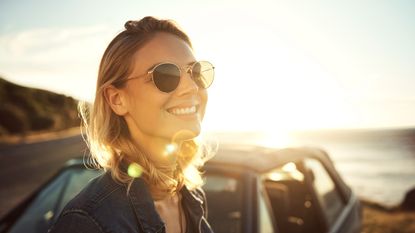 A young woman wearing sunglasses smiles with the sun shining behind her.