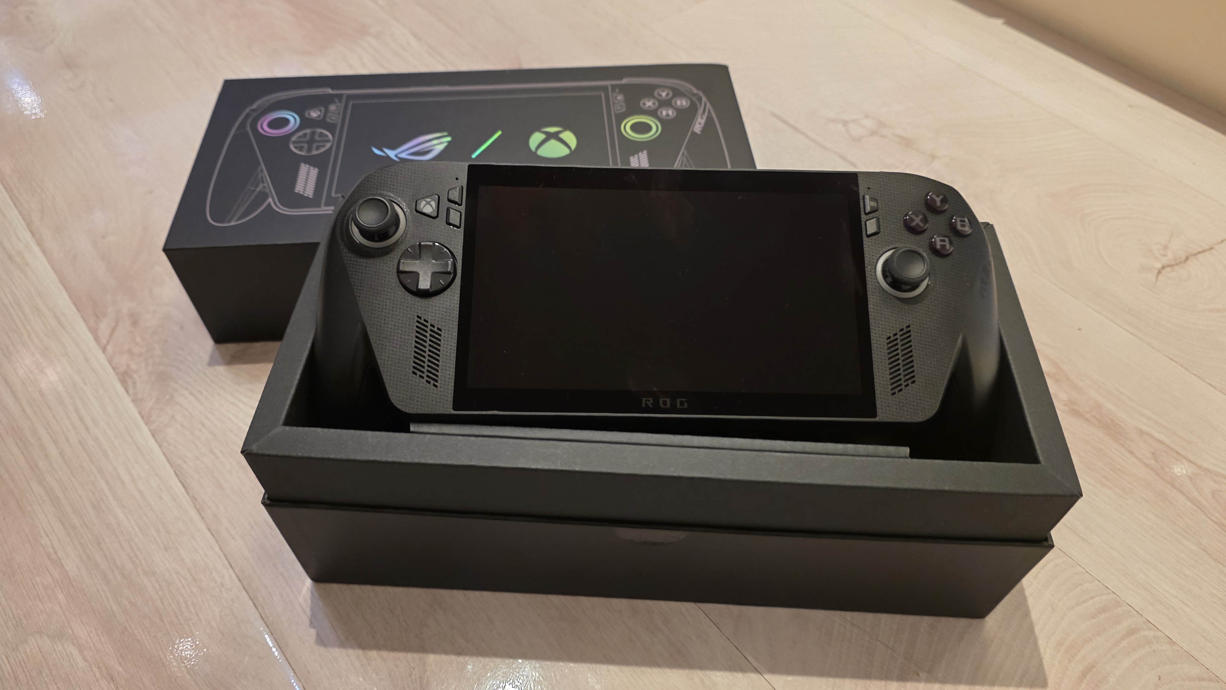 A black ROG gaming handheld console sits in an open black box on a wooden surface. The box displays controller graphics with vibrant icons.