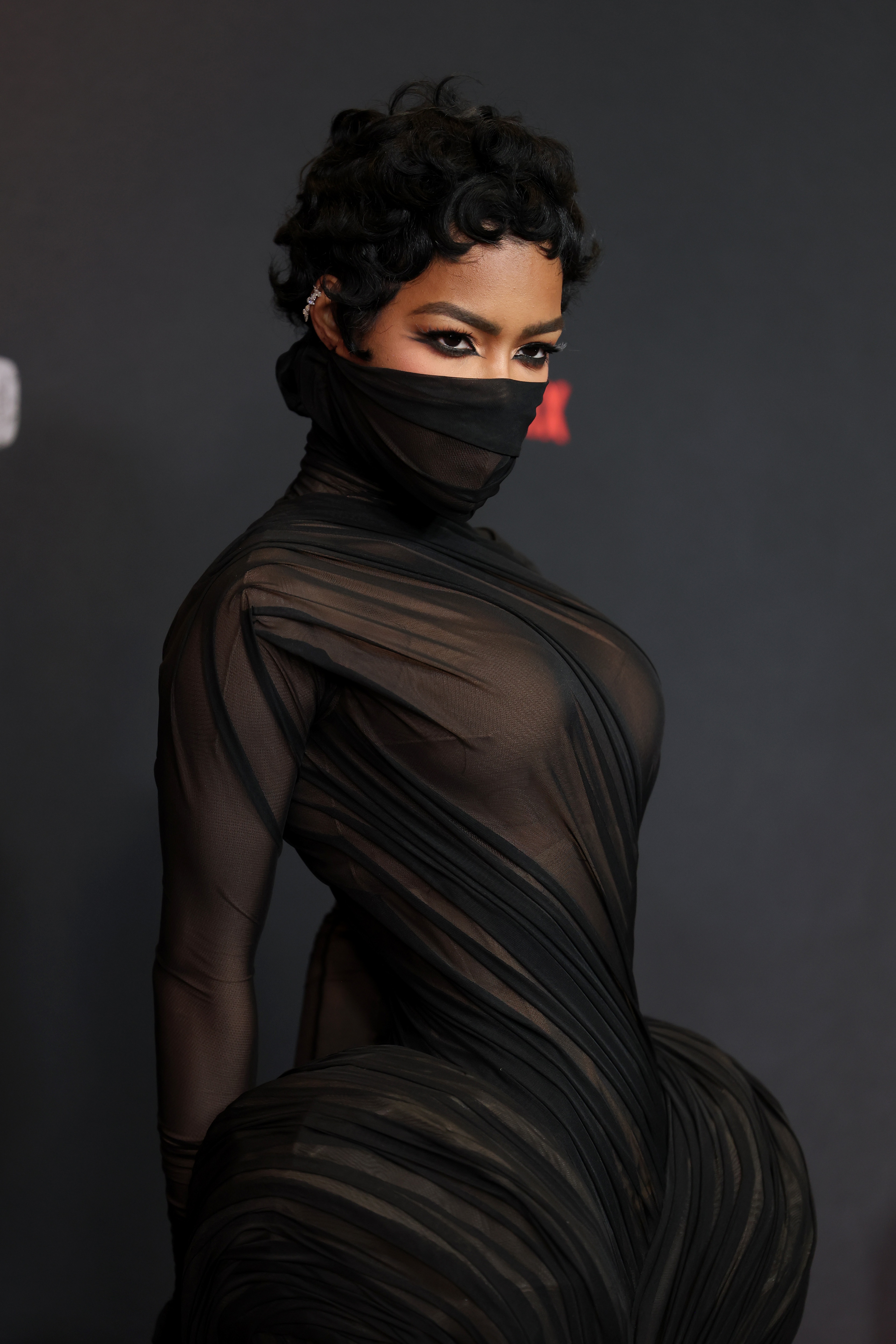 a black woman with short black hair wears a sheer gown that covers her face in front of a black background