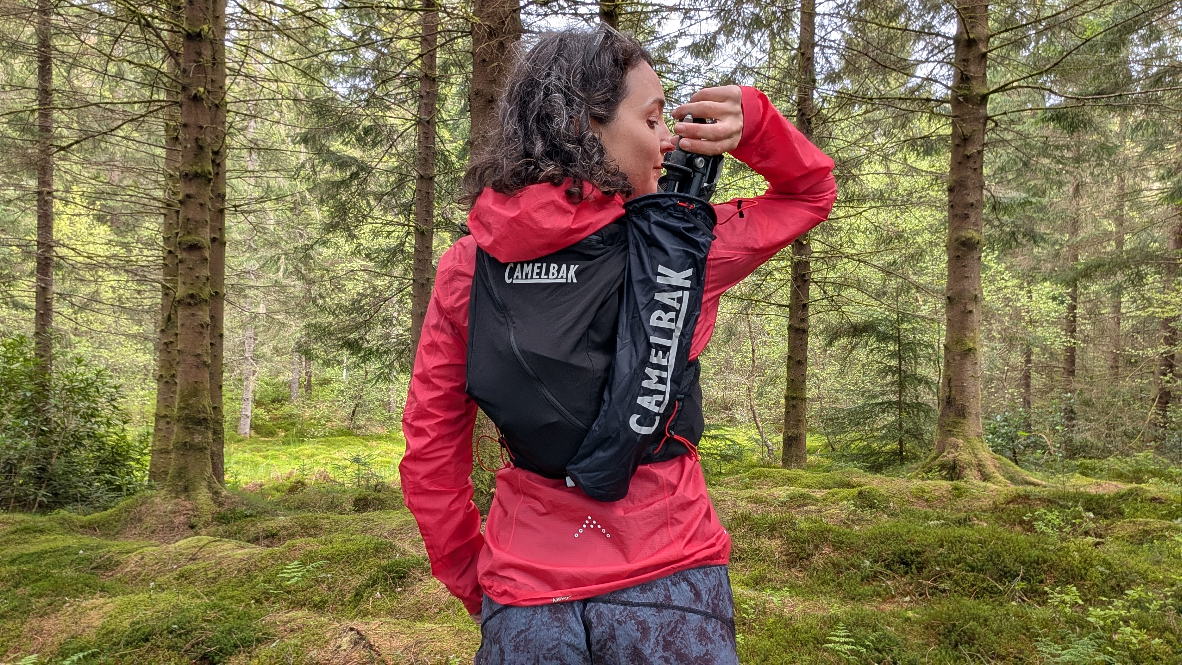 A woman trail runner in a pink jacket reaches for the poles in her black Camelbak Apex Pro running vest