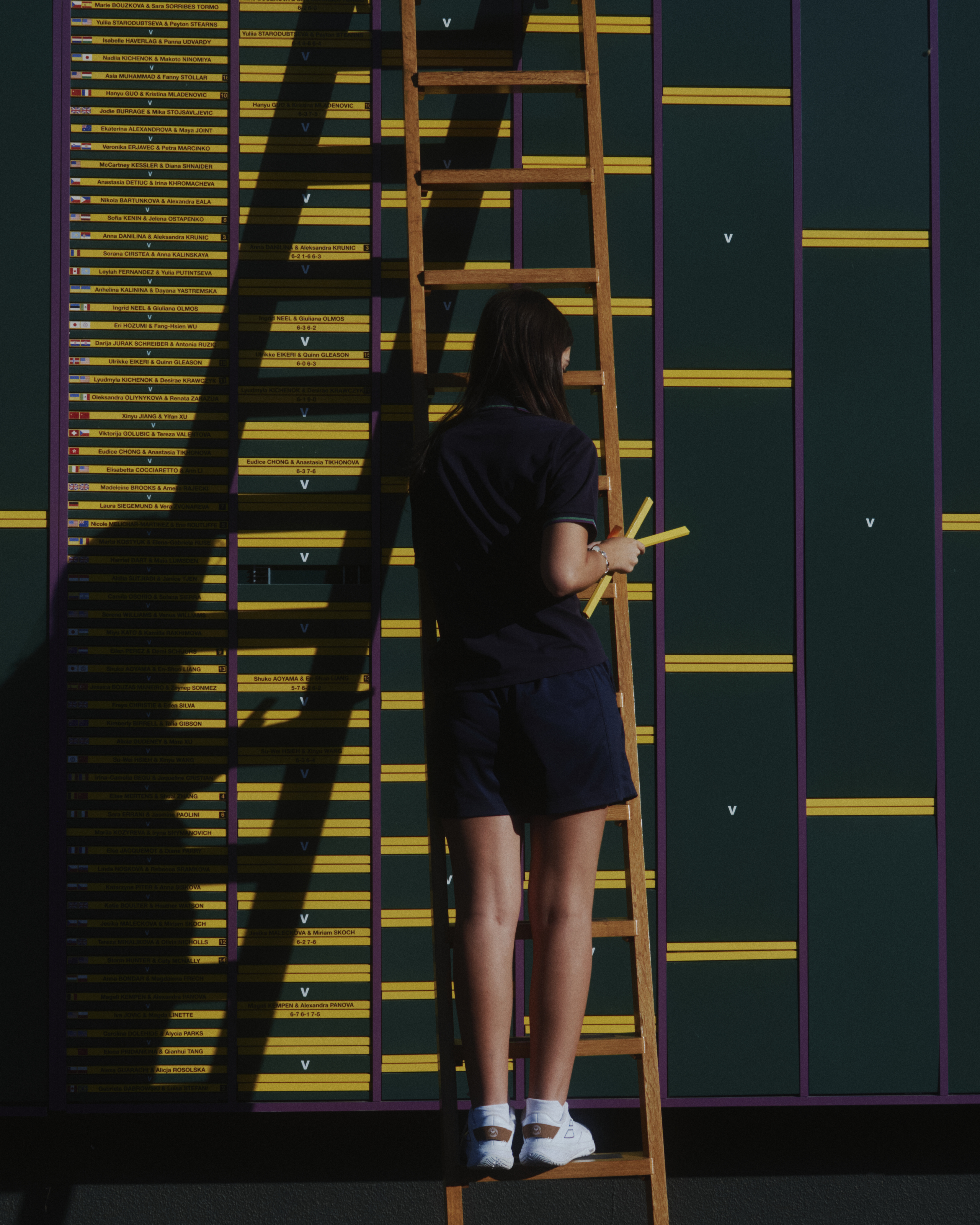 Wimbledon behind the scenes: woman up a ladder adds players' yellow name cards into slots on a green background, with purple dividing lines