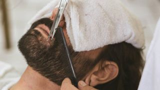 A man with a towel over his eyes, getting his beard trimmed with scissors