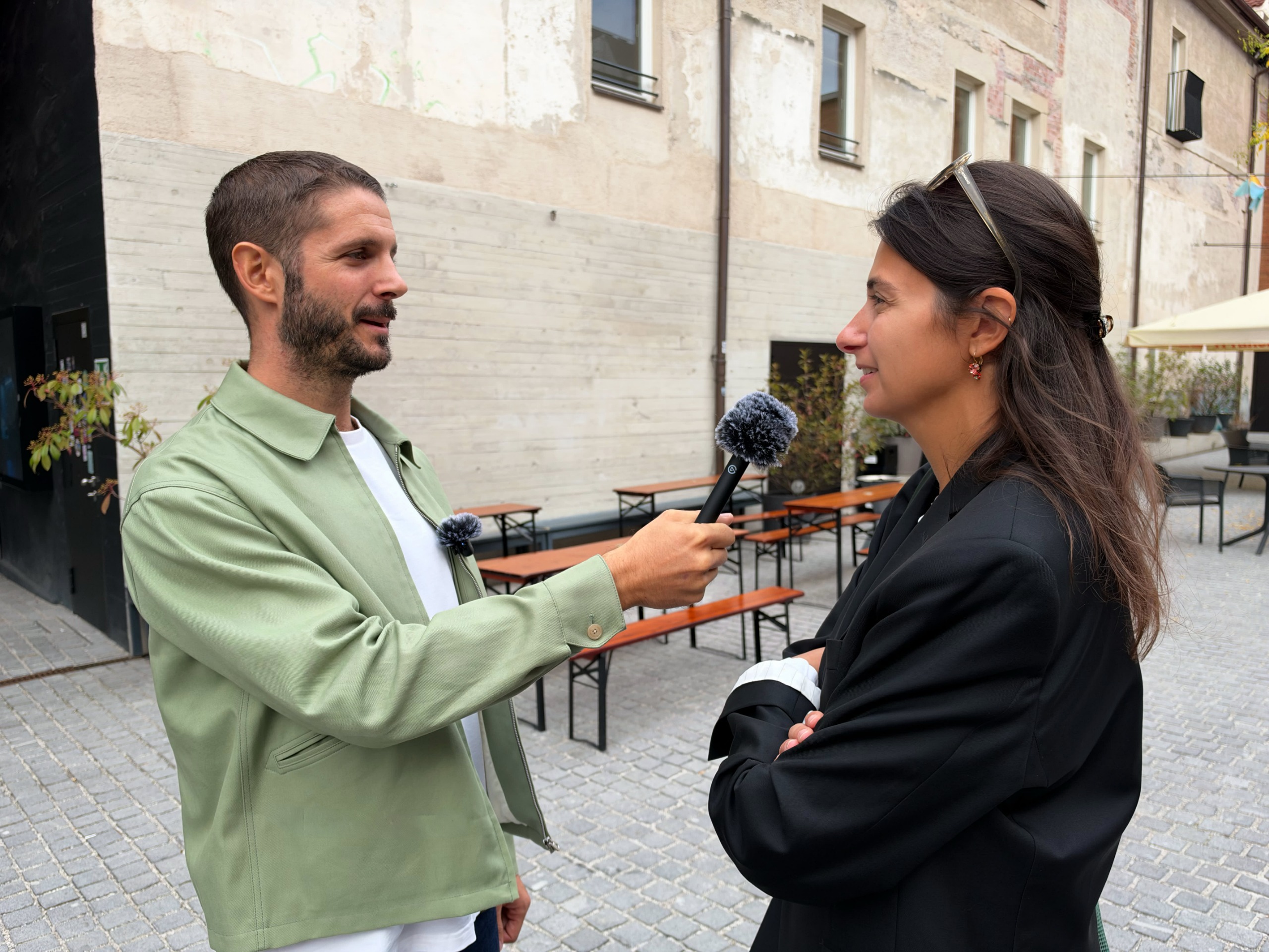 A man interviews a woman outdoors, holding a microphone as she speaks in a stone courtyard with caf&amp;eacute; benches; the scene feels relaxed and conversational.