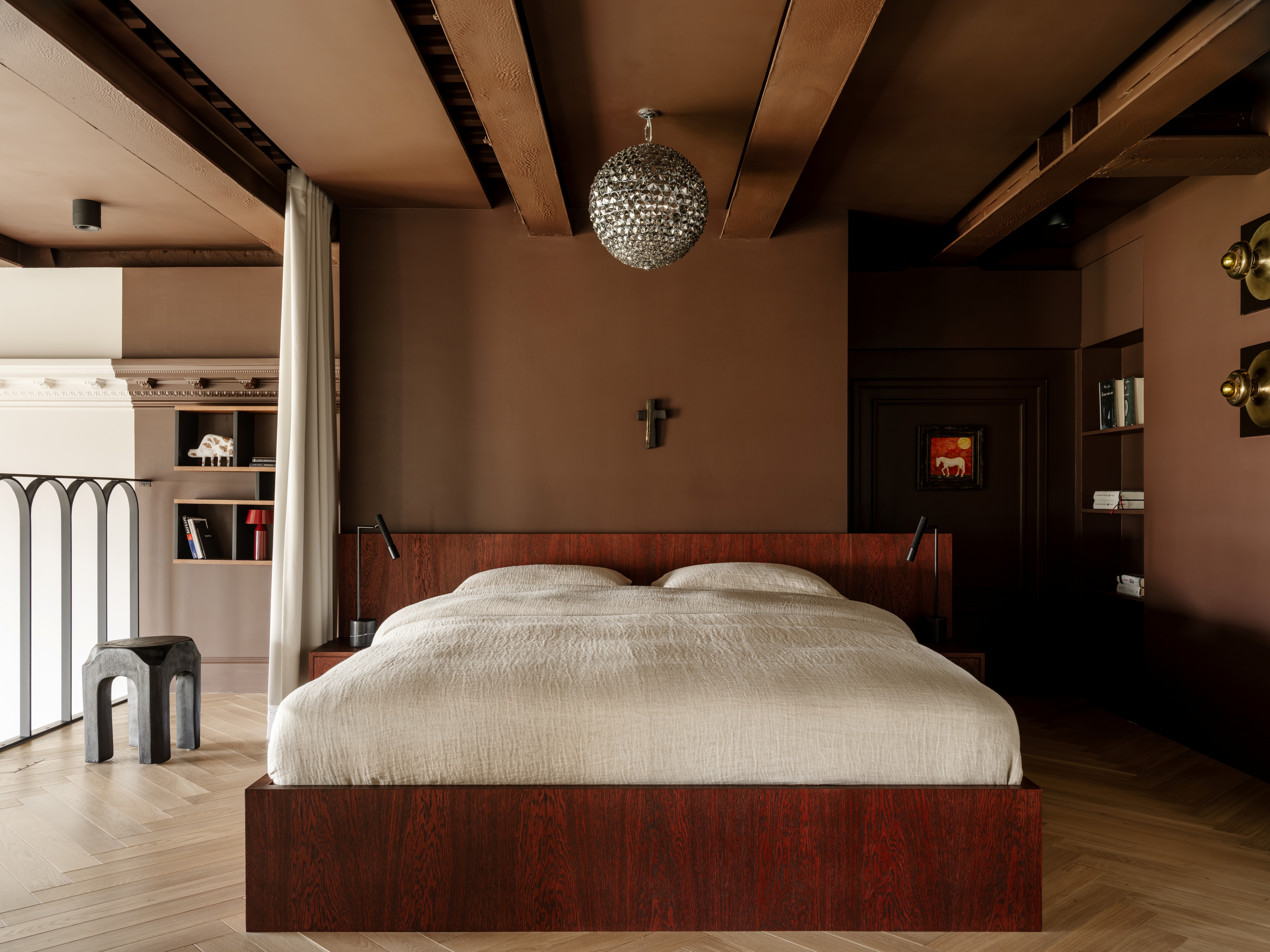 A mezzanine bedroom with a low bedframe in wood, satin beige bedsheets, sculptural furniture, brown walls, and circular sconces, alongside a centrally placed disco ball hovering above a cross.