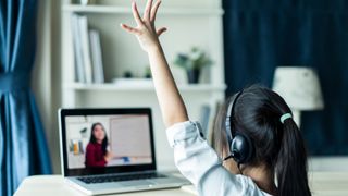 A child raises her hand while a teacher delivers a lesson online through a laptop