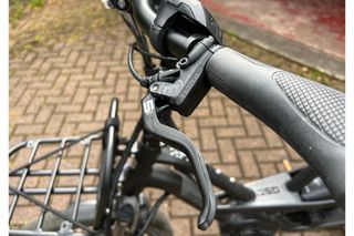 This image shows a close up of the left brake leaver and handbar grip, in the background is the rest of the bike, including front cargo frame, while the bike is stood on a brick paved driveway