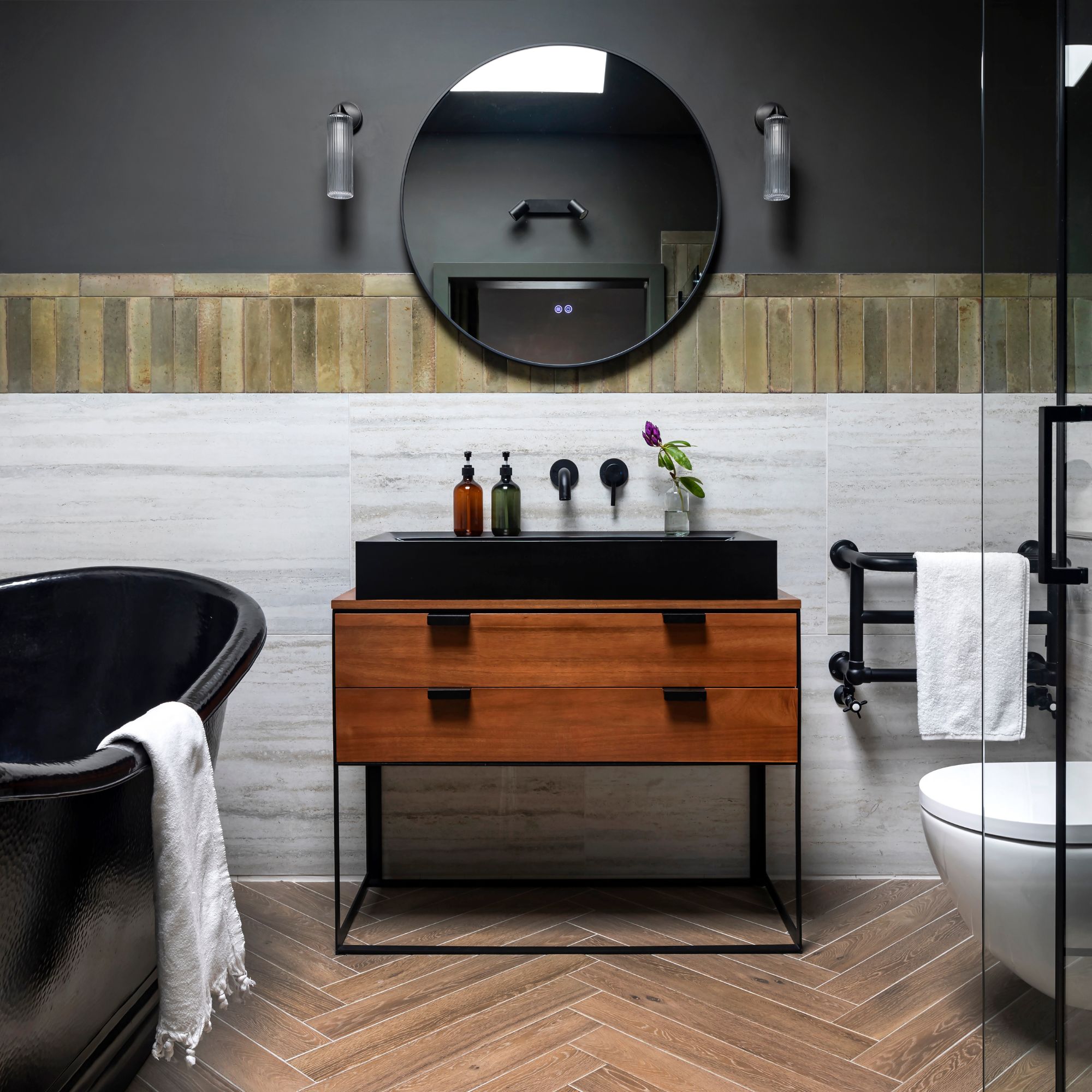 Black bathroom with wooden sink unit, black tub and accents and large round mirror
