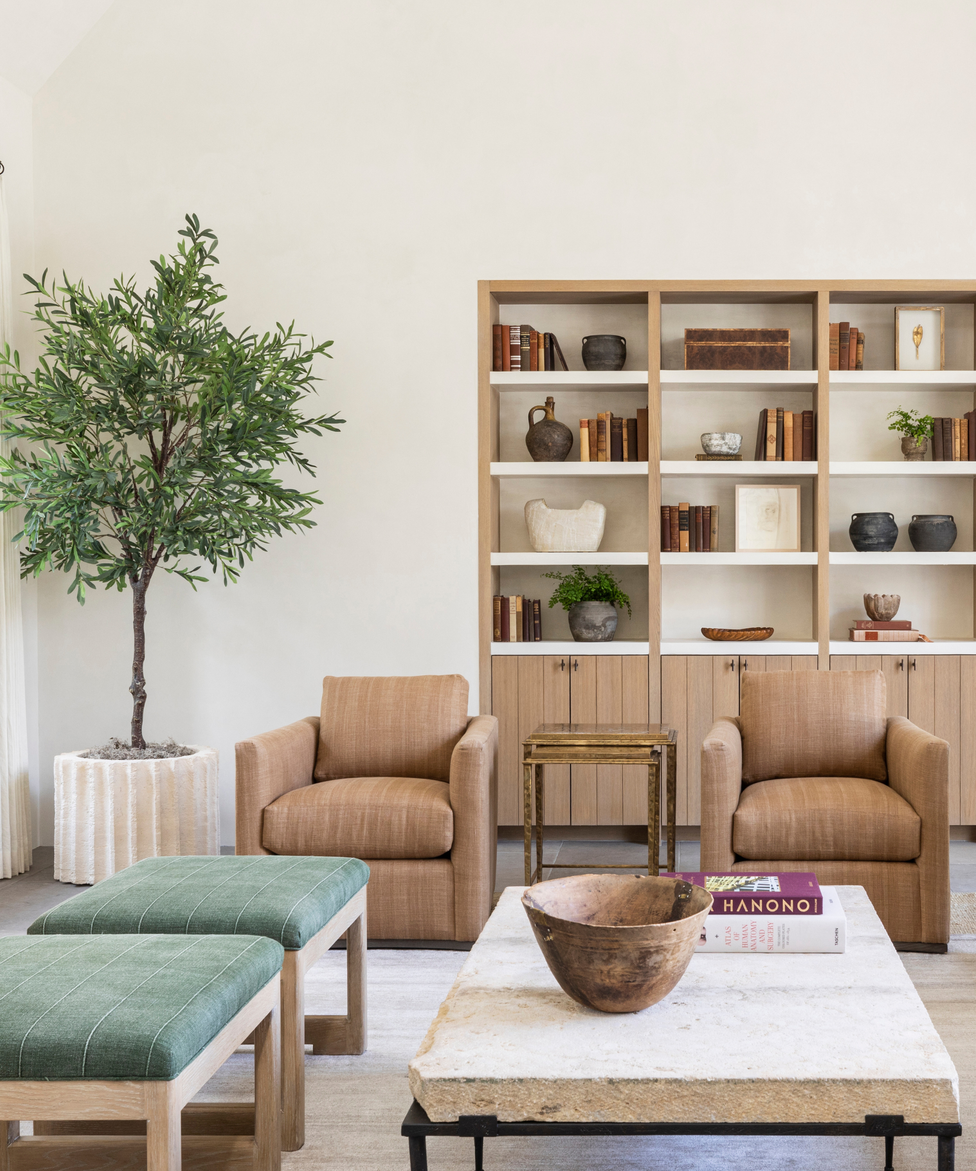A neutral-toned living room with a brown couch, a large tree, and blue ottomans