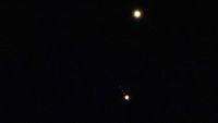 Venus is pictured shining brightly above Jupiter in a black sky. A number of small star-like objects can be seen in a line surrounding Jupiter, representing its four largest Galilean Moons.