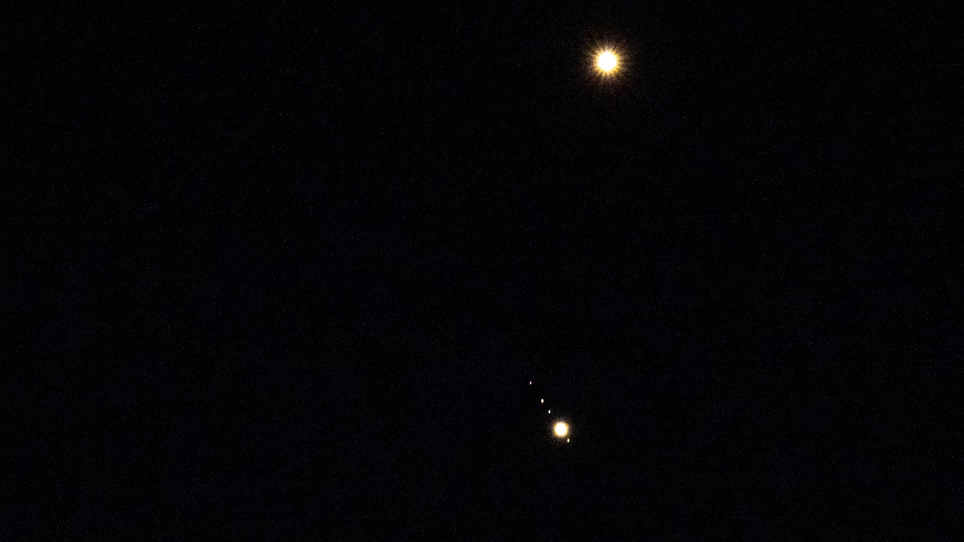 Venus is pictured shining brightly above Jupiter in a black sky. A number of small star-like objects can be seen in a line surrounding Jupiter, representing its four largest Galilean Moons.