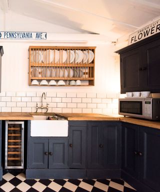 A kitchen with white tiles, dark blue cabinets, black and white checkerboard flooring, and a wooden shelf